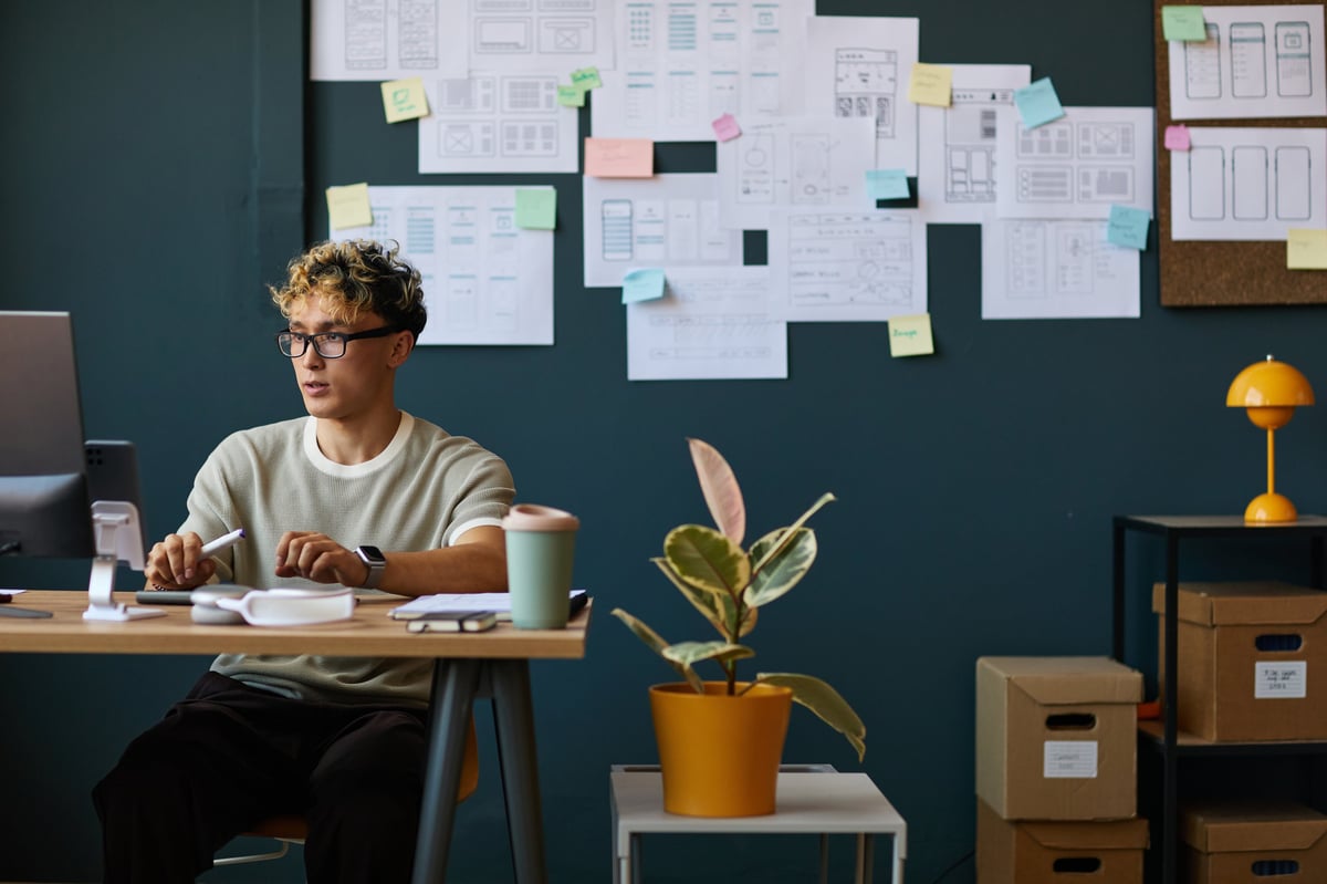 Caucasian young adult man sitting at desk working with digital tablet and stylus in modern office, design sketches and sticky notes covering wall behind, plant and boxes visible