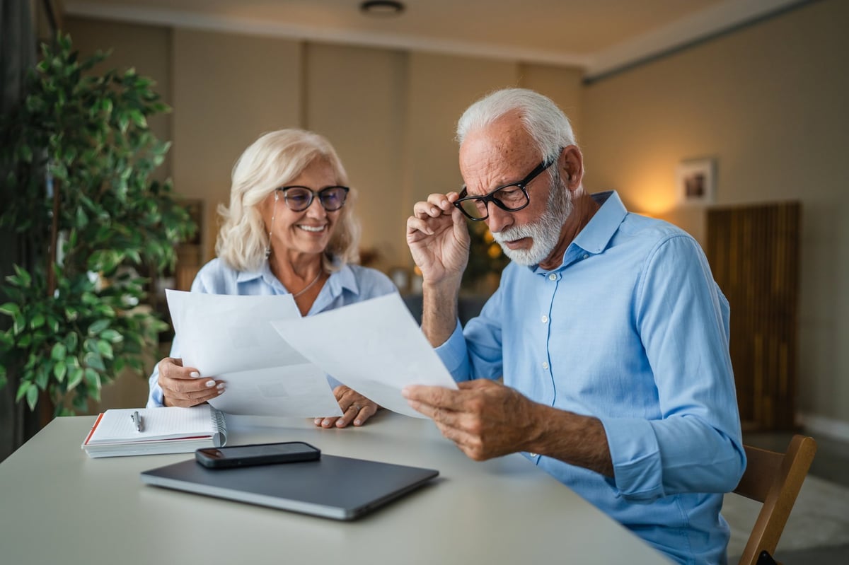 Senior couple at home smiling over financial paperwork, reviewing budgets, savings and insurance together as they plan retirement, taxes, mortgage and a secure future